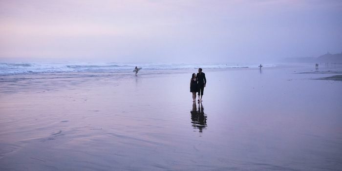 Roimata & Tristan holding hands on Muriwai Beach in Auckland at sunset, with a surfer in the distant background